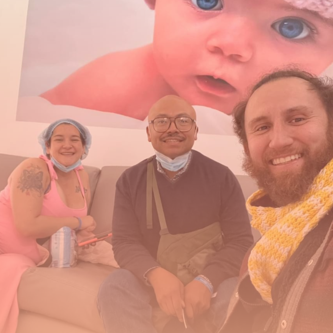 Photo of Marlon and his husband with their first surrogate, smiling together in a Colombian clinic under a large baby photo—celebrating real surrogacy stories.